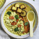 A bowl of noodle soup with tofu cubes, vegetables, sliced chilies, cilantro, and a spoon.