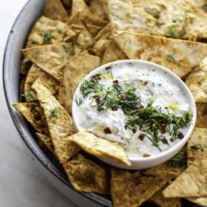 Pita chips served in a big blue bowl. A small white bowl filled with tzatziki sauce placed in it.