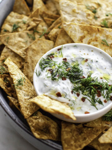 Pita chips served in a big blue bowl. A small white bowl filled with tzatziki sauce placed in it.