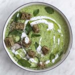 A bowl of cucumber gazpacho topped with croutons, radish and cucumber slices, and olive oil.