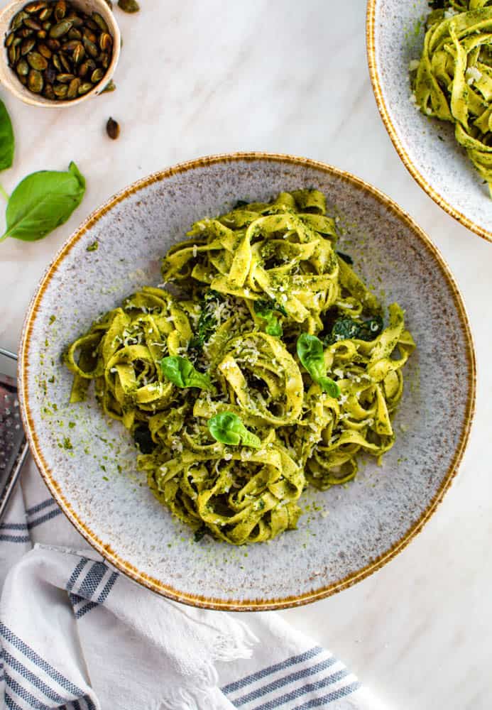 An overhead shot of a light blue bowl with pesto pasta, topped with basil leaves. A small bowl filled with pumpkin seeds next to it, alongside a white towel.