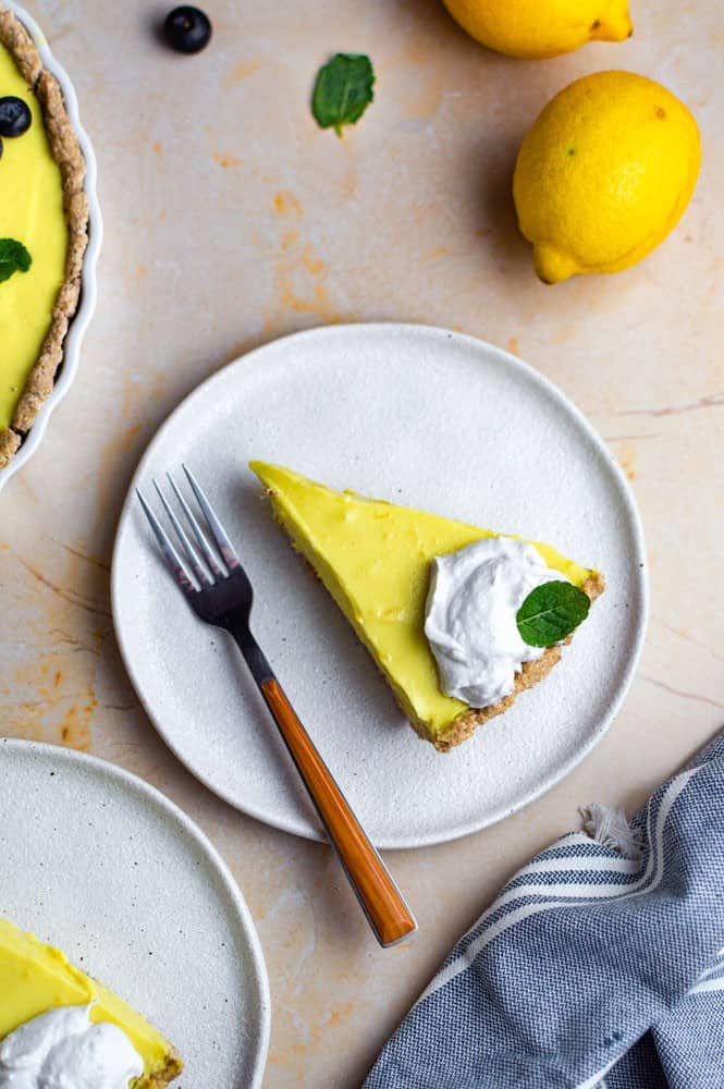 A piece of vegan lemon tart on a white plate, topped with coconut whipped cream and a mint leaf. A fork placed on the left side of the pate.