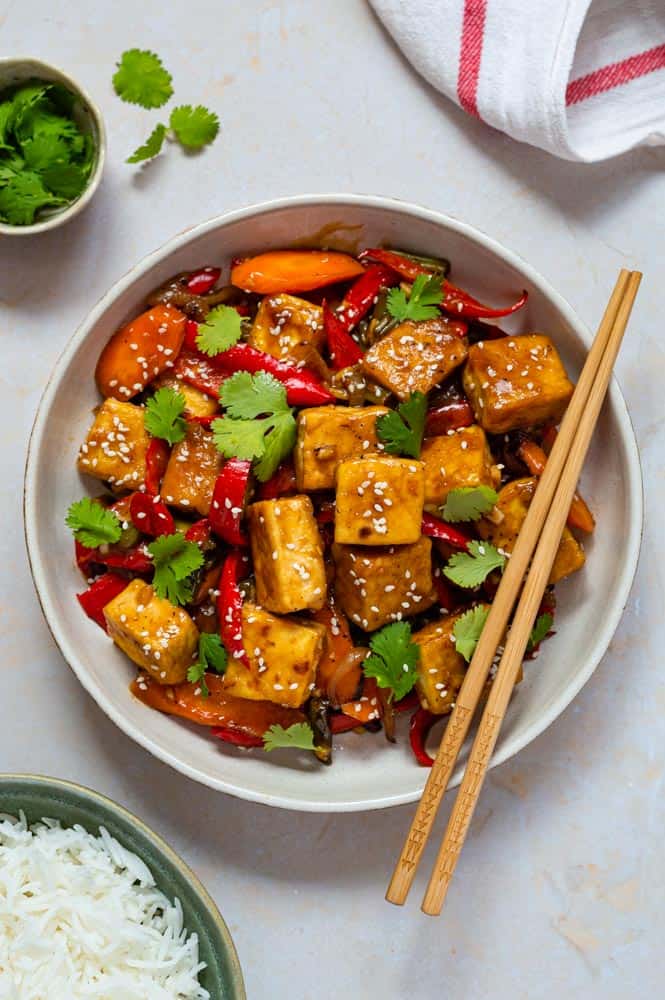 A shallow bowl of vegan stir fry with tofu. Topped with sesame seeds and coriander. Wooden chopsticks placed on the side of the plate. 