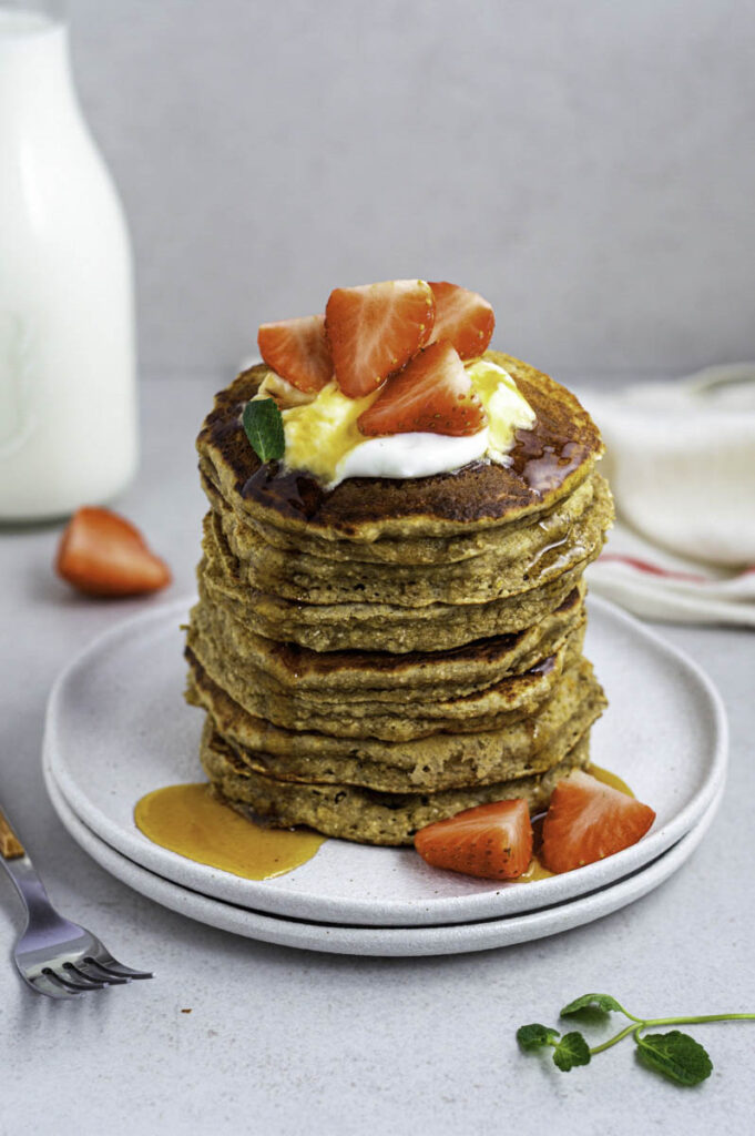A stack of banana oat pancakes on a white plate, topped with yogurt, strawberries, and honey.