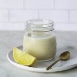 A small jar filled with white dressing placed on a white plate next to a lemon wedge and a dessert spoon.