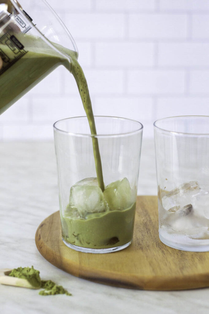 A measuring jug pouring oat milk matcha latte into a glass filled with ice cubes.