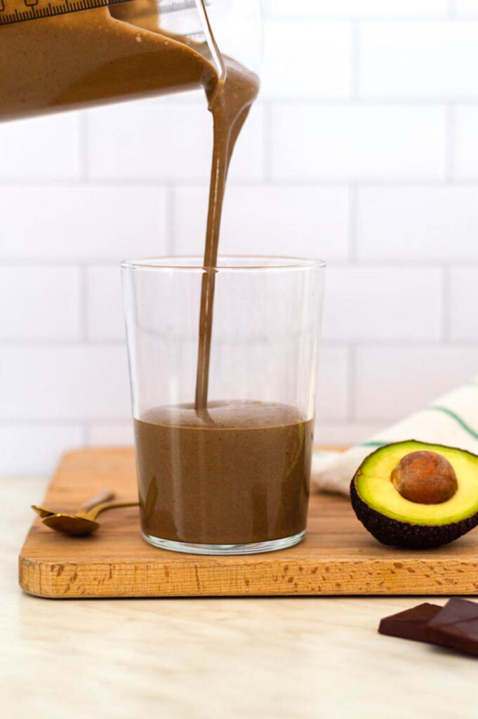 A glass placed on a wooden cutting board. A measuring jug is pouring smoothie into it. Half avocado placed on the right side of the glass.