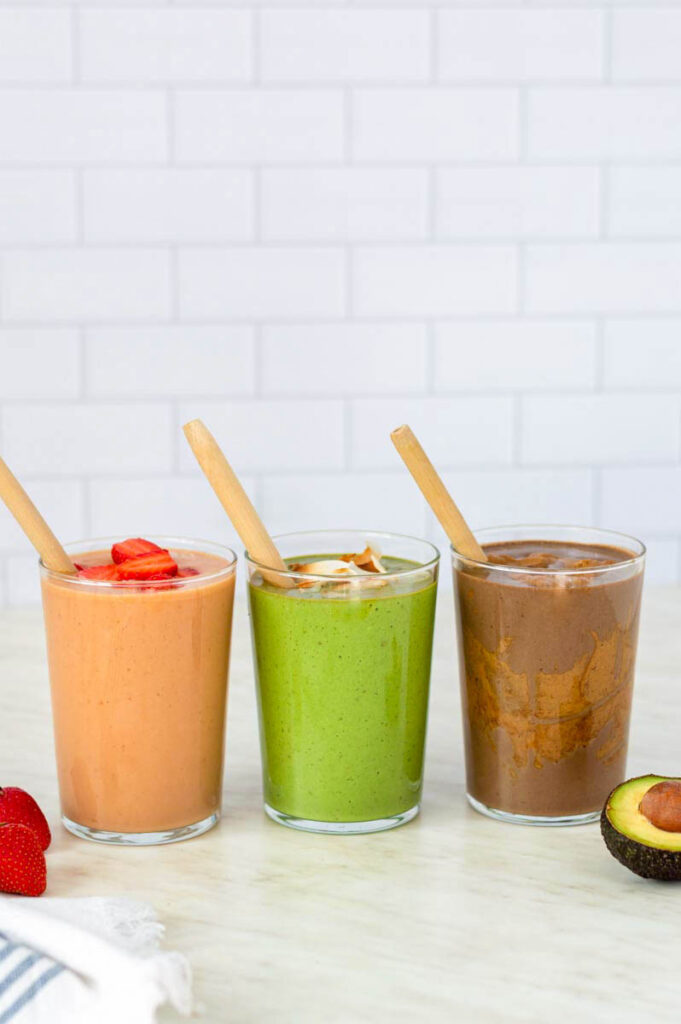 Three tall glasses placed on a kitchen countertop, filled with smoothies to the top. Wooden straws sticking out of each glass.