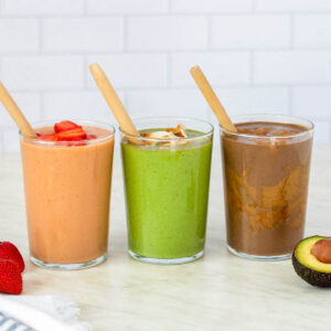 Three tall glasses placed on a kitchen countertop, filled with smoothies to the top. Wooden straws sticking out of each glass.