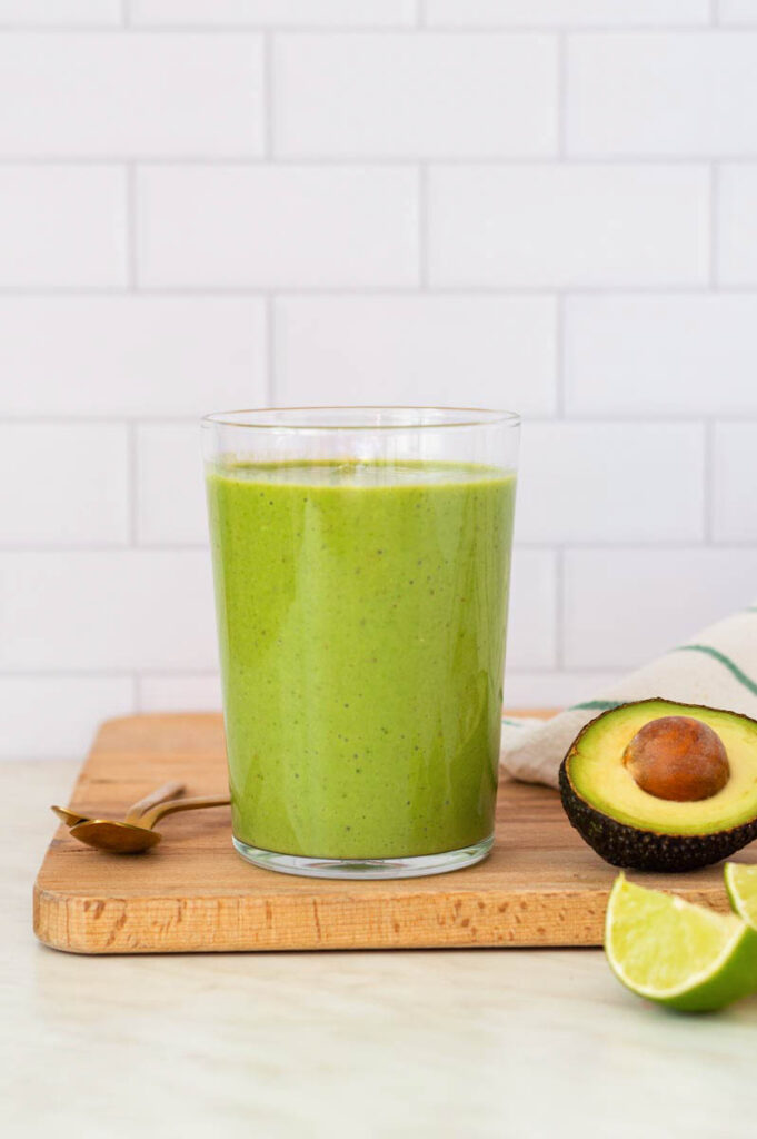 A tall glass filled with green smoothie, placed on a wooden cutting board. Half avocado and a quarter of lime placed on the right side of the glass.