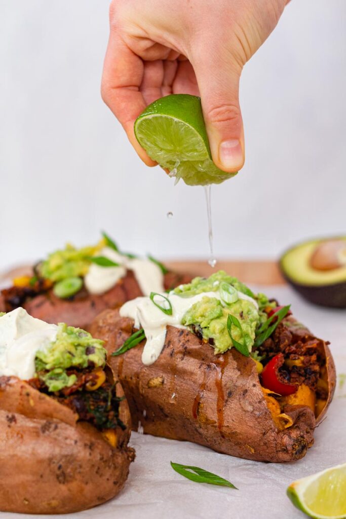 A hand is squeezing lime juice on top of a stuffed sweet potato. The sweet potato is topped with guacamole and cashew sauce.