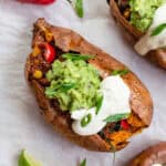 An overhead shot of roasted sweet potato, stuffed with black bean filling. The potato is topped with guacamole, cashew cream, and finely sliced green onion.