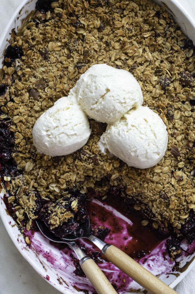 A close-up shot of blueberry crisp in a white baking dish, topped with 3 soups of ice cream