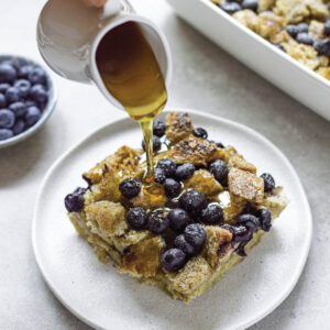 Pouring honey over a piece of blueberry French toast casserole served in a white plate.