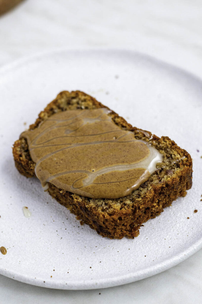 A slice of banana bread on a white plate. Topped with peanut butter and honey.