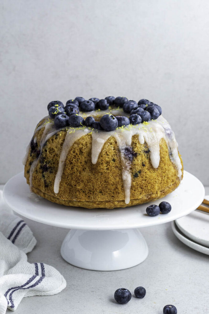 Bundt cake on a white cake stand, topped with powdered sugar glaze and fresh blueberries.