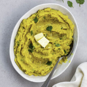 A white bowl filled with curried mashed potatoes, topped with fresh cilantro leaves and butter.
