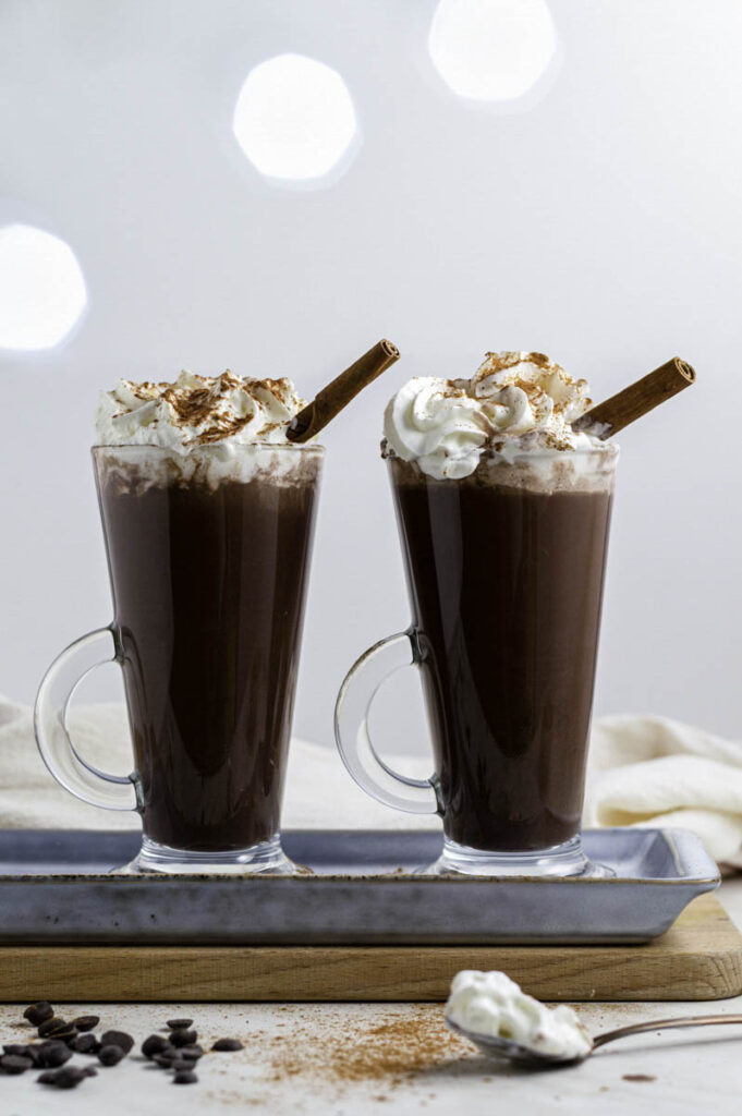 Two clear glasses filled with hot chocolate and topped with whipped cream. A cinnamon stick pointing out of each one of them.