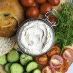 A clear jar filled with vegan cream cheese. Bagels, vegetables and fresh dill placed around it.