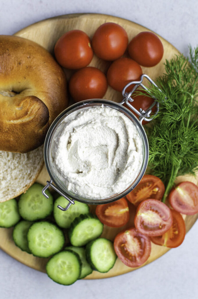 A clear jar filled with vegan cream cheese. Bagels, vegetables and fresh dill placed around it.