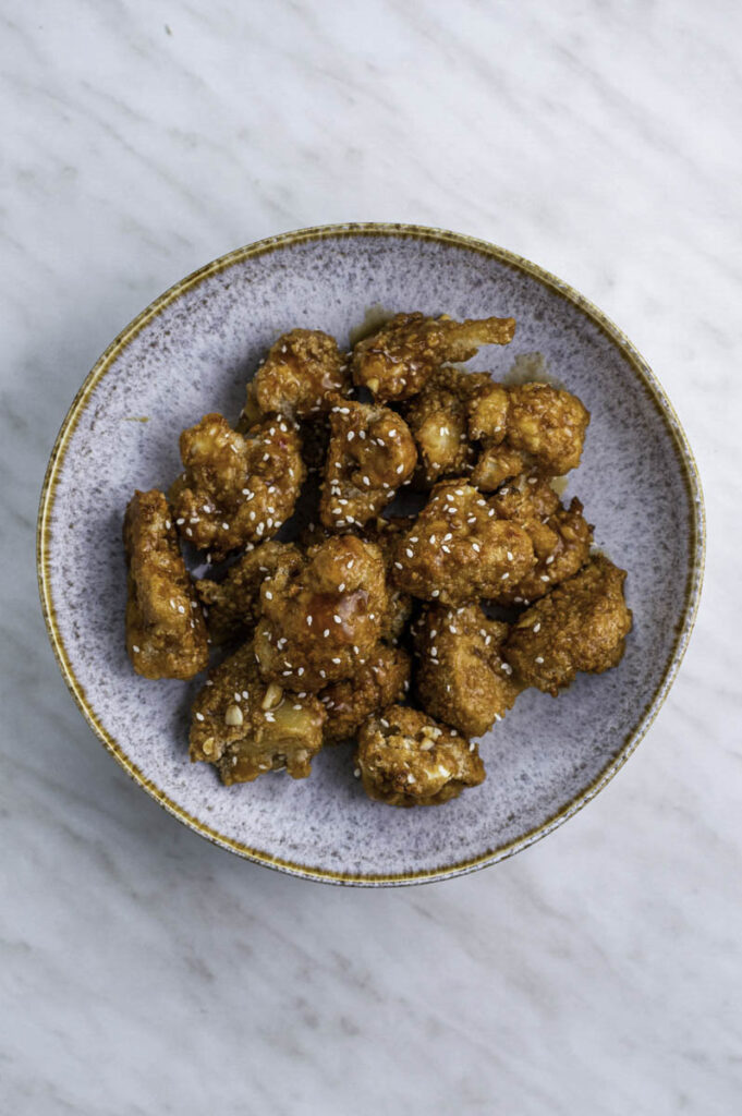 Cauliflower wings coated in teriyaki sauce in a blue bowl, and topped with sesame seeds.