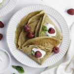 Almond milk crepes on a white plate, topped with raspeberries, yogurt, and basil.