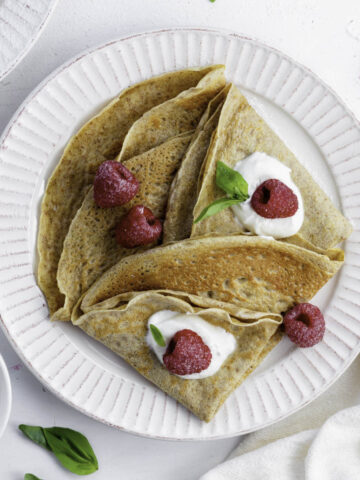 Almond milk crepes on a white plate, topped with raspeberries, yogurt, and basil.