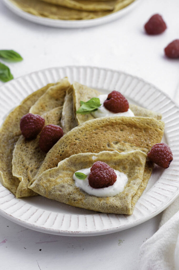 Almond milk crepes on a white plate, topped with raspberries, yogurt, and basil.