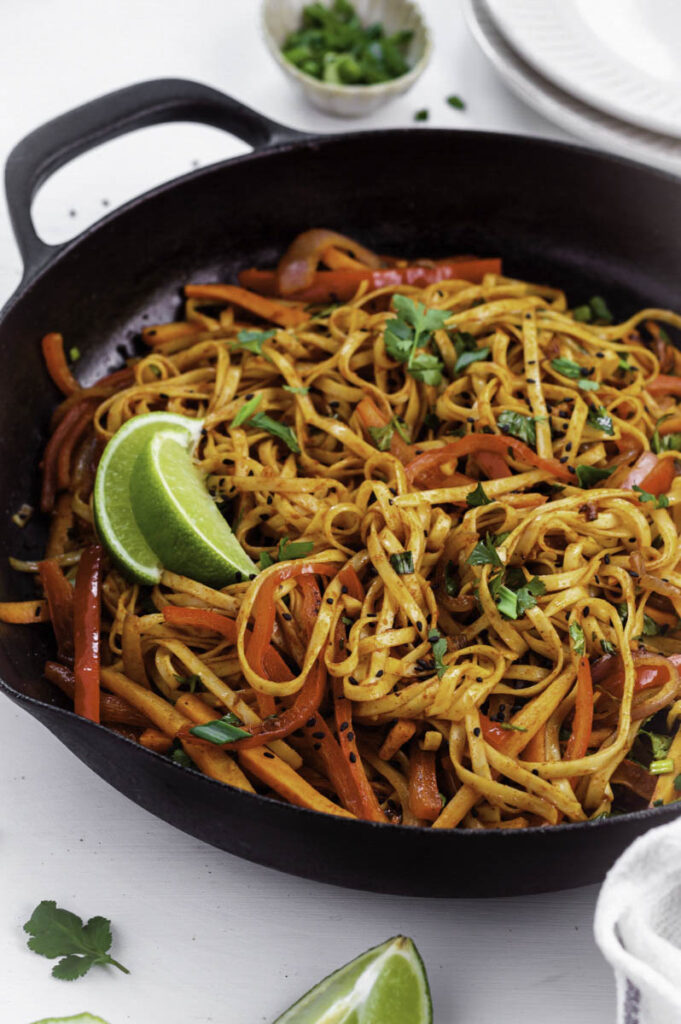 A cast-iron skillet filled with chili oil noodles, topped with fresh cilantro, and with lime slices on the side.