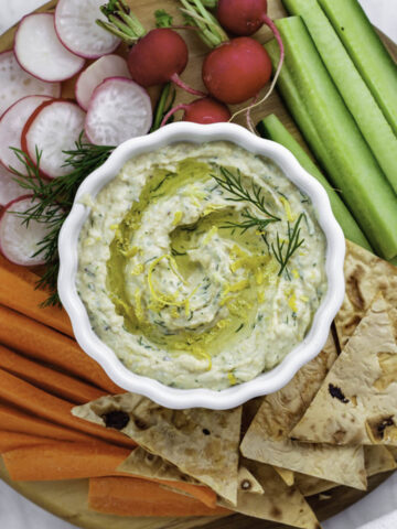 Lemon dill hummus in a white bowl served with fresh vegetable and pita chips on the side.