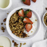 A white bowl filled with granola, strawberries, and yogurt. The bowl is placed on a marble tray.