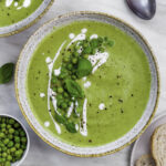 Broccoli pea soup served in a blue bowl, topped with peas, coconut milk and fresh basil leaves.