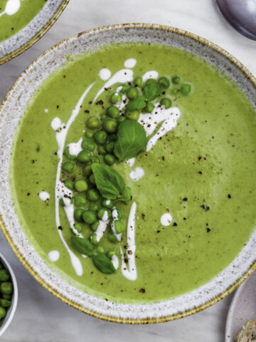 Broccoli pea soup served in a blue bowl, topped with peas, coconut milk and fresh basil leaves.