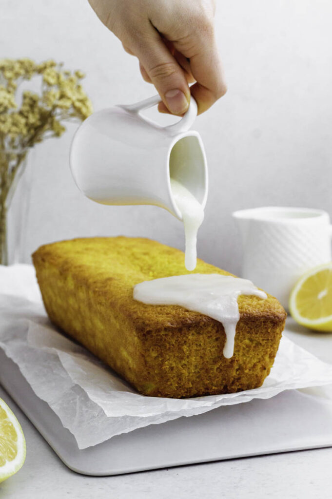 A hand pouring powdered sugar glaze from a small white jug over lemon cake.
