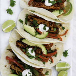 Four flour tortillas filled with tofu fajitas, and served on a white plate with parchment paper.