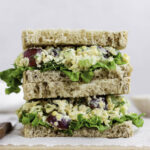 A stack of two vegan chicken salad sandwiches on a wooden cutting board.
