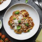 A white plate filled with tomato ravioli topped with grated parmesan cheese and fresh basil leaves.