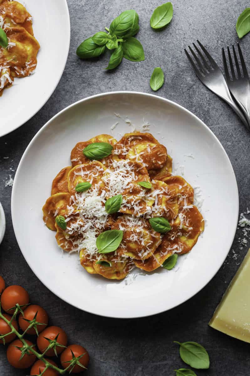 A white plate filled with Ravioli Pomodoro topped with grated parmesan cheese and fresh basil leaves.