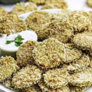 Baked zucchini crisps on a white plate with a little bowl full of ranch dressing.
