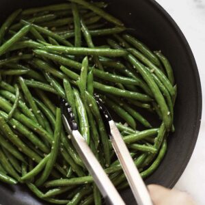 Garlic parmesan green beans cooking in a black pan. Stirring the green beans using pasta tongs.