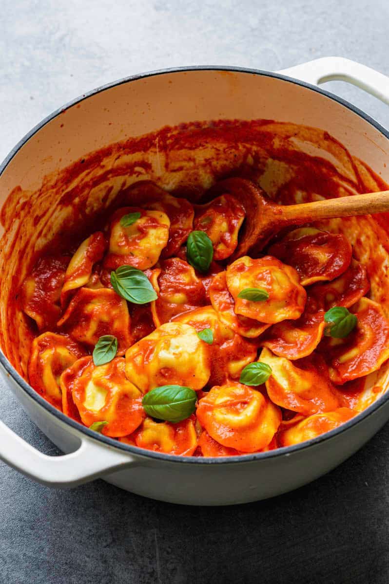 Ravioli with Pomodoro sauce in a white Dutch oven with a wooden spoon in it, topped with fresh basil leaves.