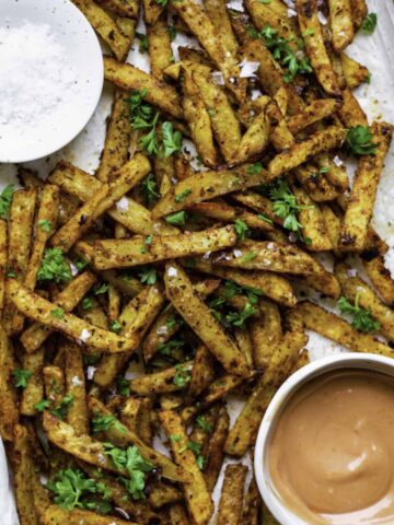 Five guys Cajun fries served on a baking sheet layered with parchment paper. They're topped with flaky sea salt and chopped parsley.