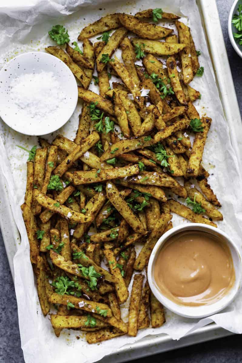 Five guys Cajun fries served on a baking sheet layered with parchment paper.  One bowl white filled with sauce placed on the lower right side of the baking sheet, and one white bowl filled with salt placed on the upper left side.