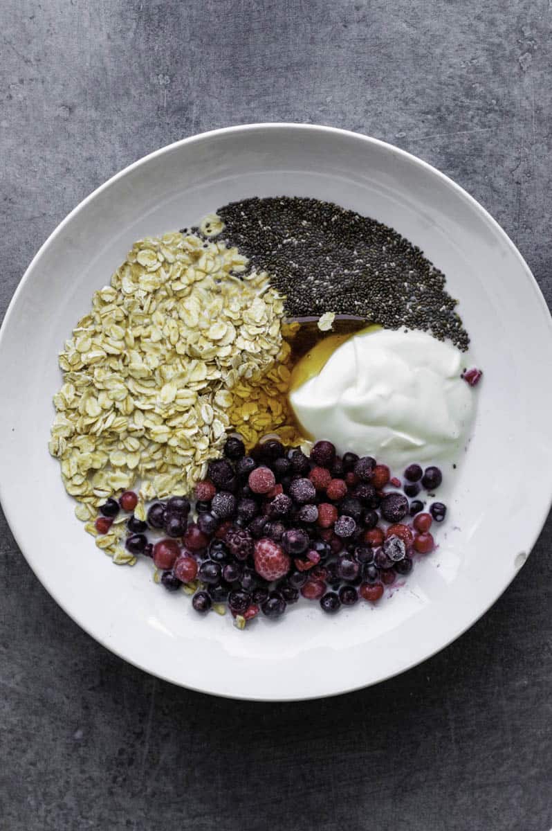 Oats, frozen berries, yogurt, chia seeds, pinch of salt, and maple syrup added to white bowl. 