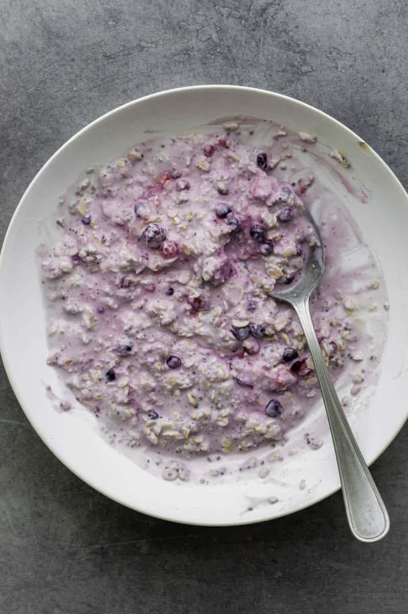 Oats, frozen berries, yogurt, chia seeds, pinch of salt, and maple syrup mixed with water in a white bowl with a silver spoon in it.