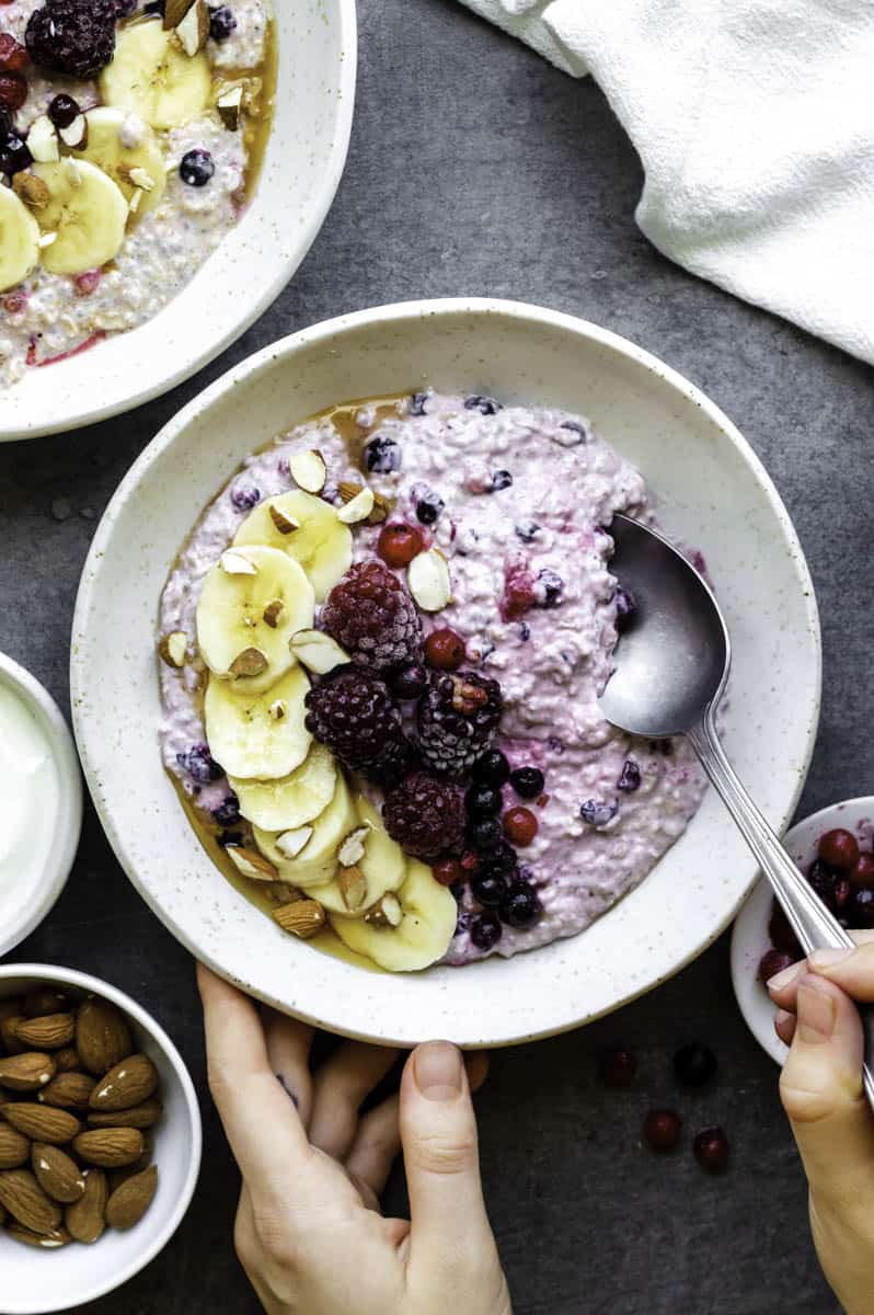 One hand holding a white bowl filled with overnight oats topped with a sliced banana, blackberries, and almonds. The other hand is dipping a spoon in the bowl. 