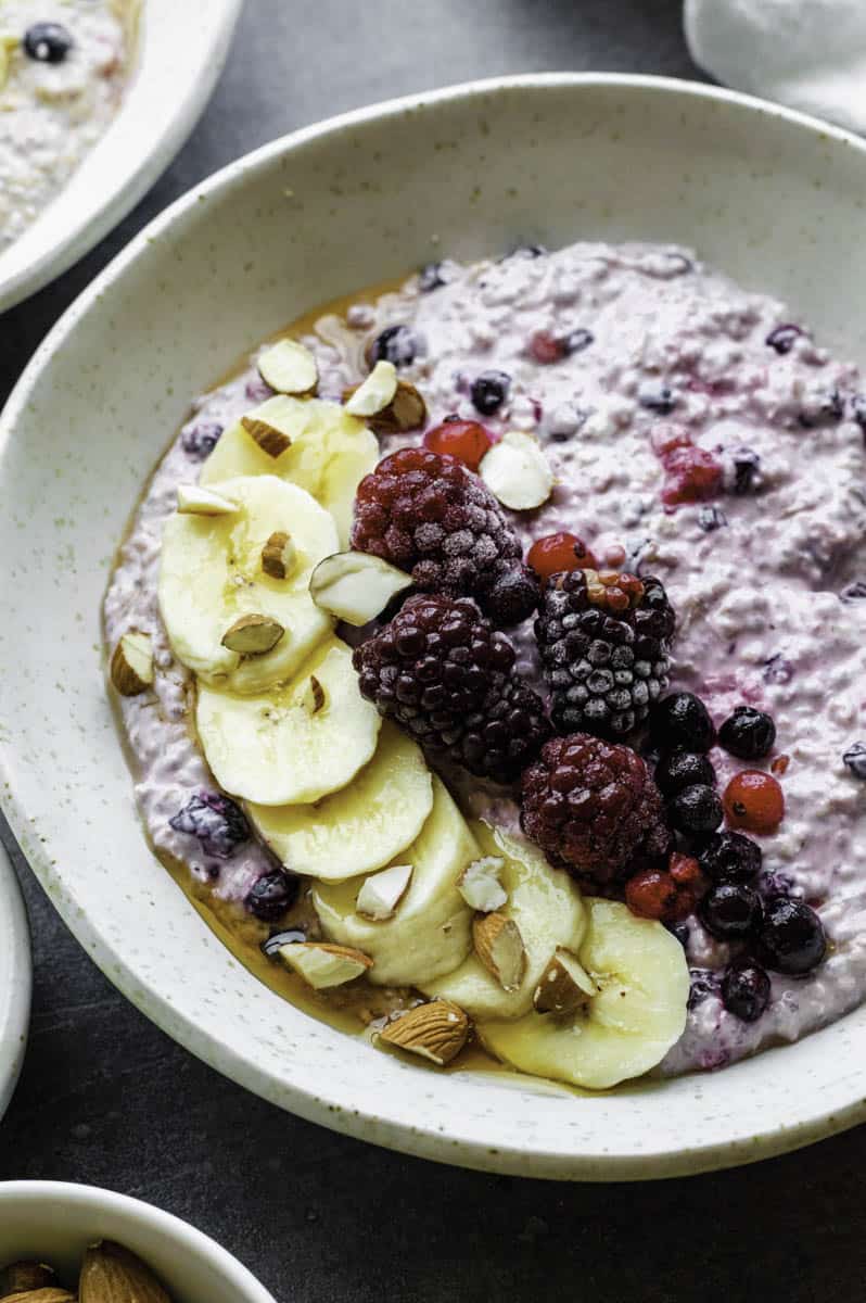 A white bowl filled with overnight oats with frozen fruit topped with a sliced banana, blackberries, and crushed almonds.