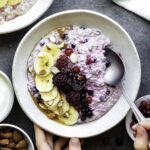 One hand holding a white bowl filled with overnight oats topped with a sliced banana, blackberries, and almonds. The other hand is dipping a spoon in the bowl.