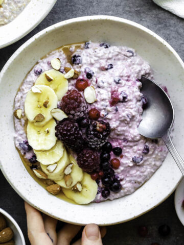 One hand holding a white bowl filled with overnight oats topped with a sliced banana, blackberries, and almonds. The other hand is dipping a spoon in the bowl.