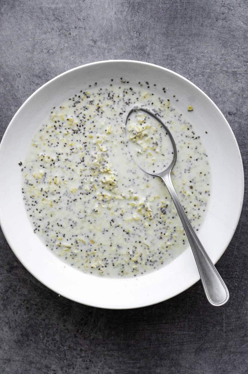 Oats, yogurt, chia seeds, pinch of salt, and maple syrup mixed with water in a white bowl with a silver spoon in it.